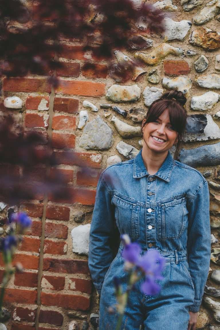 Naturpathic nutritionist Nikki leans against a stone and brick wall smiling widly. She's wearing a denim jumpsuits and part of the camera is covered with bright blue flowers to frame Nikki.